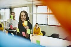 Two business women and a man in an office putting sticky notes on a window. Primary colors: yellow and blue.